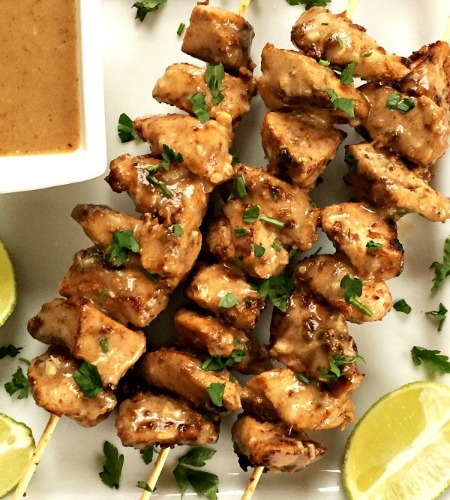 3 chicken satay skewers on a white plate with lime wedges and a bowl of peanut butter sauce