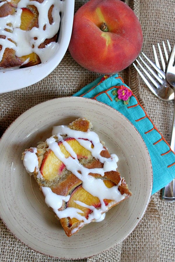 overhead shot of slice of fresh peach cake on a beige plate set on top of turquoise napkin with forks and a fresh peach in the background