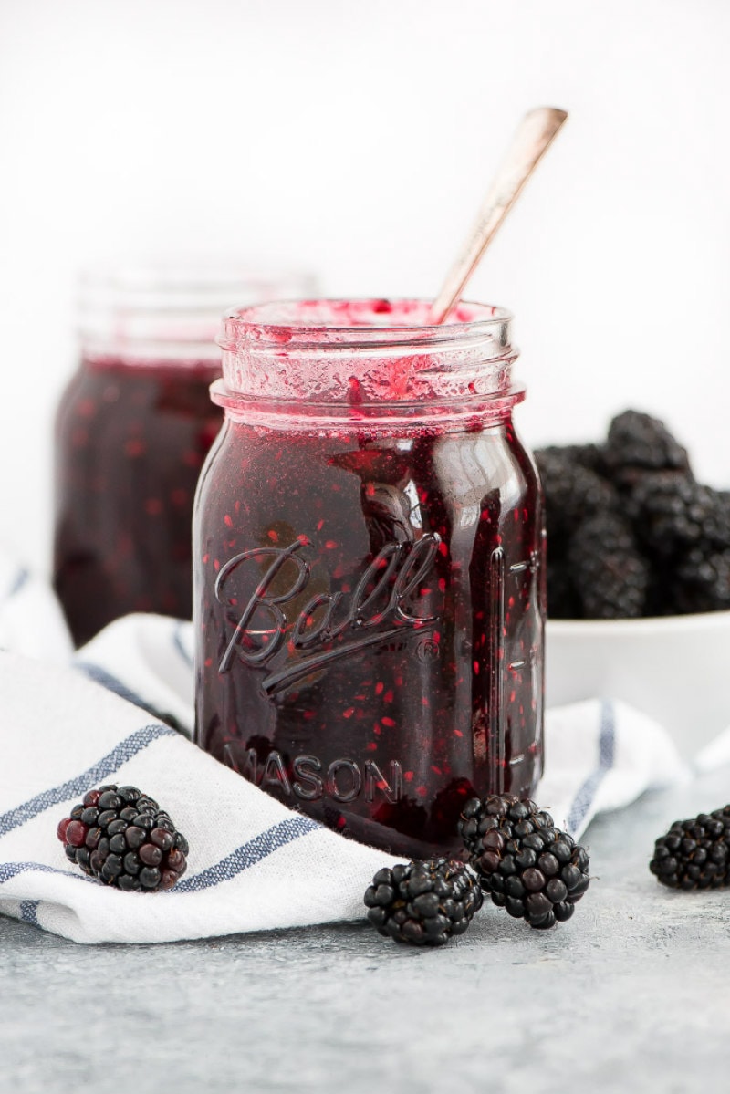 Jar of Blackberry Freezer Jam with a spoon in it, white and blue towel underneath, and fresh blackberries scattered around too