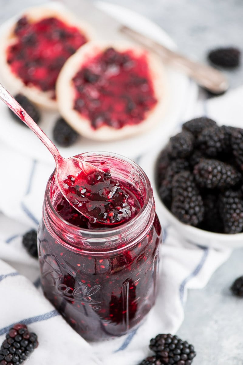 spooning out Blackberry Freezer Jam out of a jar. English muffins with jam in the background. Fresh blackberries displayed too